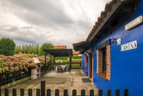 Casina Casa de color azul con un porche y jardín de flores, bajo cielo nublado.